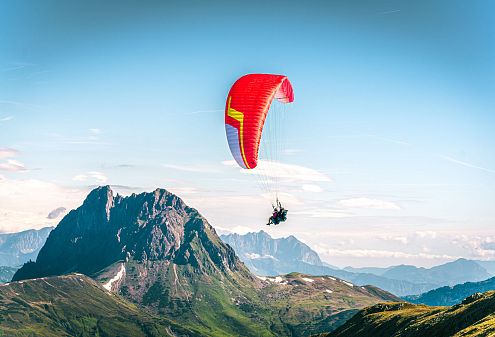 Ein Paraglider mit rotem Schirm schwebt über einer grünen Gebirgslandschaft. Im Hintergrund sind hohe, felsige Berge und ein blauer Himmel mit ein paar Wolken zu sehen.