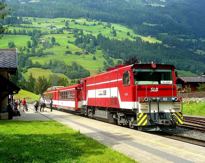 Ein roter Zug fährt durch eine malerische grüne Landschaft mit Hügeln. Menschen warten auf dem Bahnsteig. Im Hintergrund sind Bäume und ein bewölkter Himmel zu sehen.