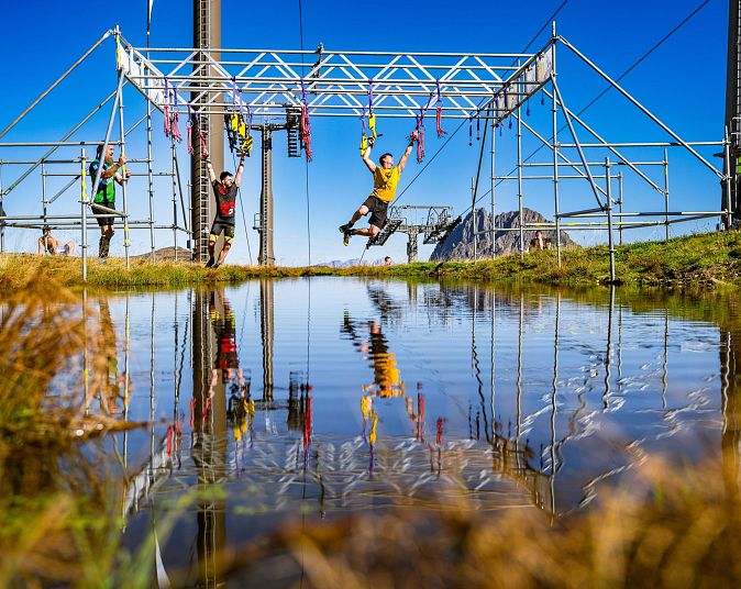Teilnehmer eines Hindernislaufs schwingen an einem Klettergerüst über einen Wassergraben, bei strahlendem Sonnenschein unter blauem Himmel.