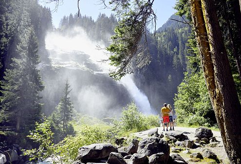 Ein Paar steht auf einem Felsen vorm Krimmler Wasserfall inmitten eines grünen Waldgebiets. Sonnenlicht fällt durch die Bäume, und Nebel steigt vom Wasser auf.