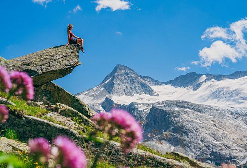 Žena sedí na skalním výběžku s velkolepým výhledem na zasněženou horu na pozadí, kolem kvetou květiny a obloha je jasná a modrá.
