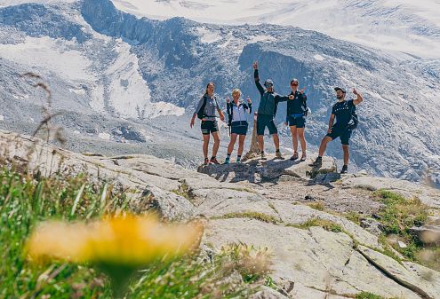 Groep wandelaars loopt over een bergpad met besneeuwde toppen op de achtergrond, onder een heldere blauwe lucht met enkele wolken.