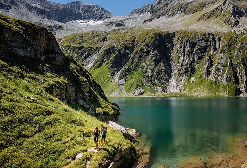 Twee wandelaars bij een schilderachtig bergmeer omringd door groene heuvels en bergen met blauwe hemel.