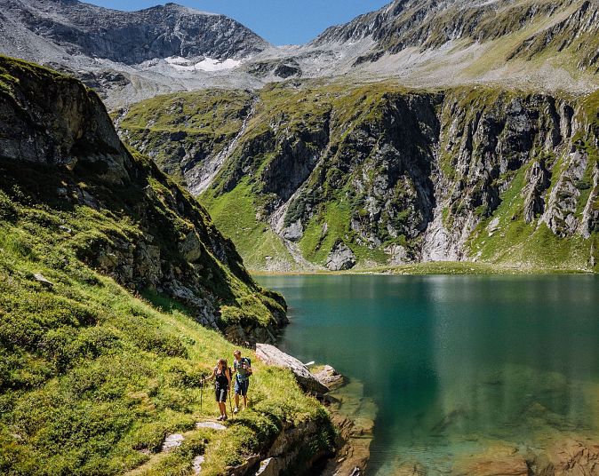 Dva turisté kráčejí po stezce vedle tyrkysového horského jezera, obklopeného strmými zelenými svahy a zasněženými vrcholky hor pod modrou oblohou.