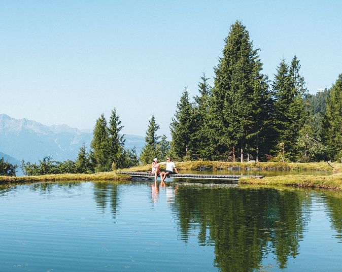 Ein ruhiger Bergsee reflektiert den klaren Himmel und die umliegenden Bäume. Im Hintergrund sind hohe Berge zu sehen. Am Ufer sitzen zwei Menschen auf einem Steg.