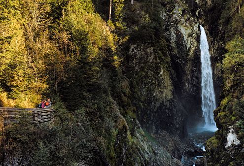 Vysoký vodopád v lese, obklopený strmými skalami a bujnou zelení, zatímco pár sleduje scenérii z vyhlídky nalevo.