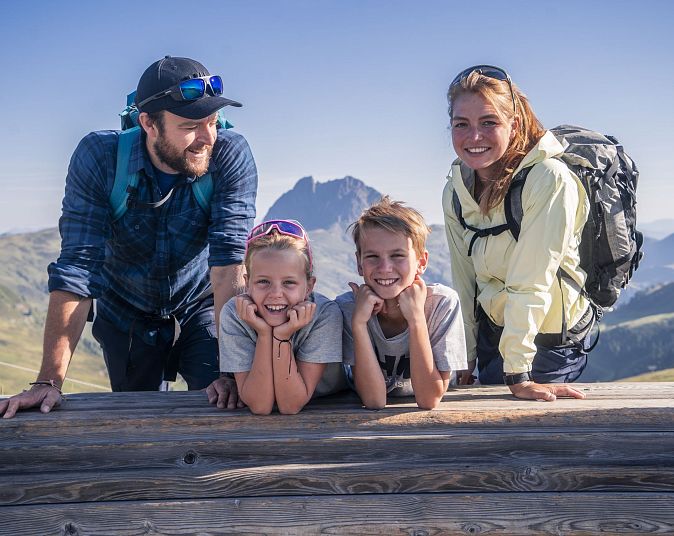 Eine Familie posiert lächelnd auf einer Berghütte vor atemberaubender Alpenlandschaft. Der Himmel ist klar, und alle tragen Freizeitkleidung und Rucksäcke.