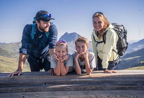 Eine Familie posiert lächelnd auf einer Berghütte vor atemberaubender Alpenlandschaft. Der Himmel ist klar, und alle tragen Freizeitkleidung und Rucksäcke.