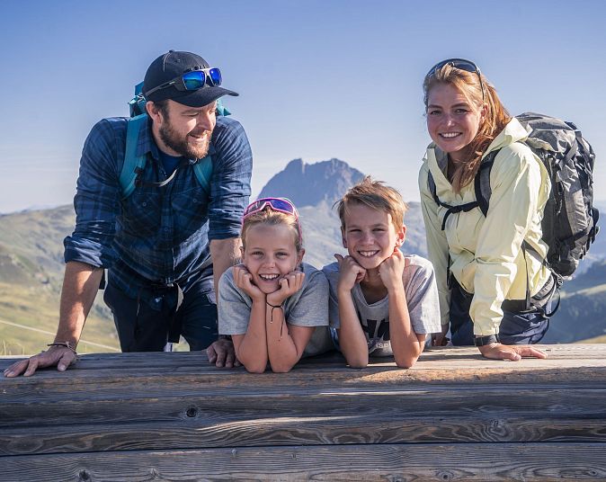 Eine Familie posiert lächelnd auf einer Berghütte vor atemberaubender Alpenlandschaft. Der Himmel ist klar, und alle tragen Freizeitkleidung und Rucksäcke.