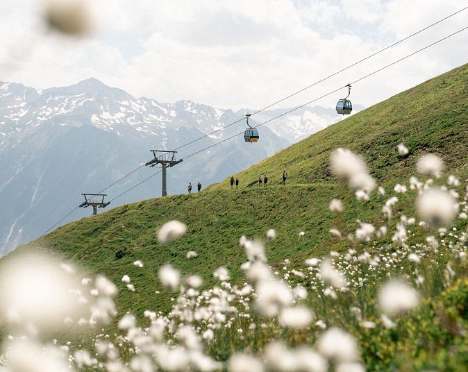 Een schilderachtig alpenlandschap met een groene heuvel bedekt met bloemen, kabelbaan en bergen op de achtergrond onder een bewolkte hemel.