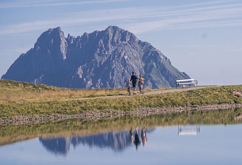 Ein Paar wandert auf einem Pfad an einem ruhigen See vorbei, der die umliegenden Berge spiegelt. Der Himmel ist klar und die Landschaft wirkt friedlich und einladend.