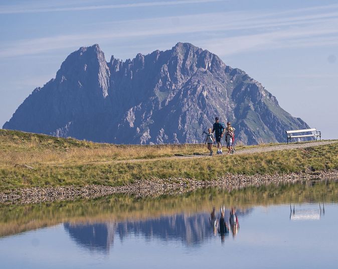 Ein Paar wandert auf einem Pfad an einem ruhigen See vorbei, der die umliegenden Berge spiegelt. Der Himmel ist klar und die Landschaft wirkt friedlich und einladend.