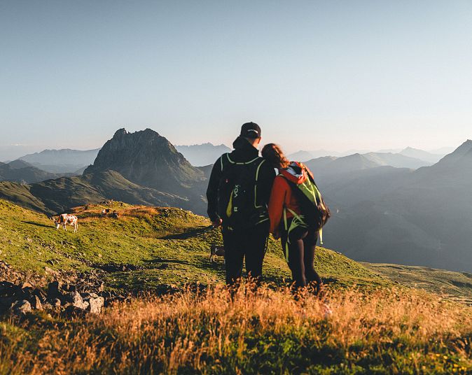 Ein Paar steht auf einem Bergpfad mit grünen Wiesen und blickt auf eine majestätische Berglandschaft. Der Himmel ist klar und die Sonne scheint sanft auf die Szenerie.