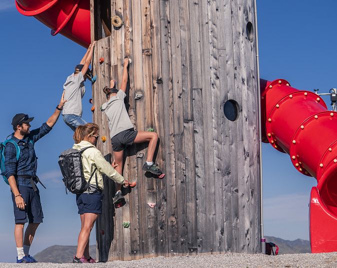 Berggipfel mit mehreren Gebäuden und einer Kletterwand, umgeben von grasbewachsenen Flächen. Menschen genießen die Aussicht, während Wolken die umliegenden Täler bedecken.