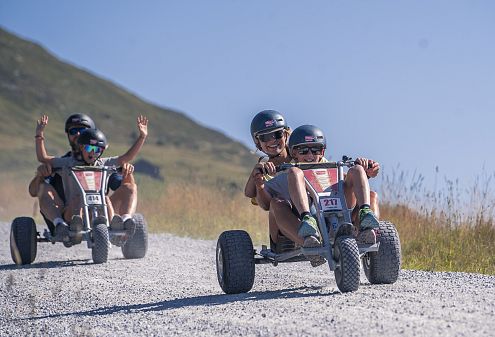 Drei Personen fahren an einem sonnigen Tag mit Mountaincarts auf einem Schotterweg in der Wildkogel-Arena. Im Hintergrund sind hohe Berge und ein blauer Himmel zu sehen.