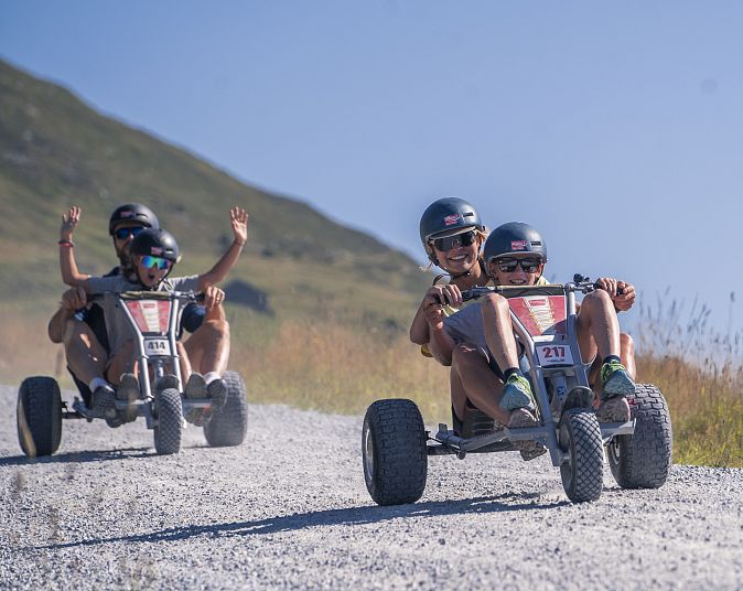 Drei Personen fahren an einem sonnigen Tag mit Mountaincarts auf einem Schotterweg in der Wildkogel-Arena. Im Hintergrund sind hohe Berge und ein blauer Himmel zu sehen.