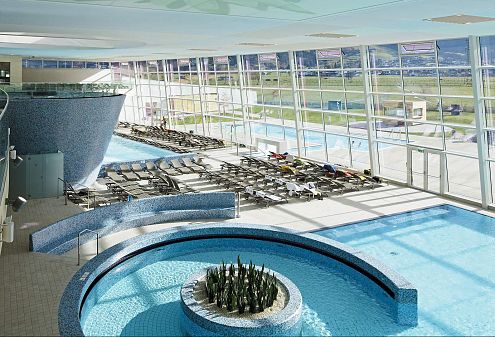 Innenaufnahme eines modernen Hallenbads mit großen Fenstern, Liegestühlen und einem runden Schwimmbecken im Vordergrund. Helle, klare Gestaltung.