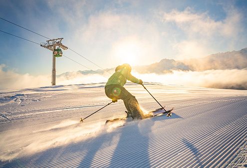 Skifahrer fährt bei strahlendem Sonnenschein auf frisch präparierter Piste bergab. Im Hintergrund sind Seilbahn und schneebedeckte Berge unter klarem Himmel zu sehen.