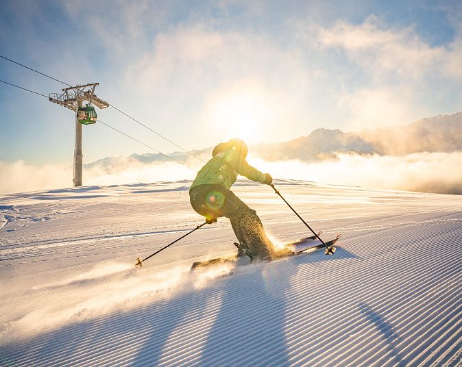 Skifahrer fährt bei strahlendem Sonnenschein auf frisch präparierter Piste bergab. Im Hintergrund sind Seilbahn und schneebedeckte Berge unter klarem Himmel zu sehen.