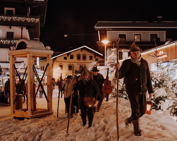 Winterliche Straßenszene in einem verschneiten Dorf bei Nacht. Menschen mit Wanderstäben und Laternen schlendern durch den beleuchteten Ort, umgeben von traditionellen Gebäuden.