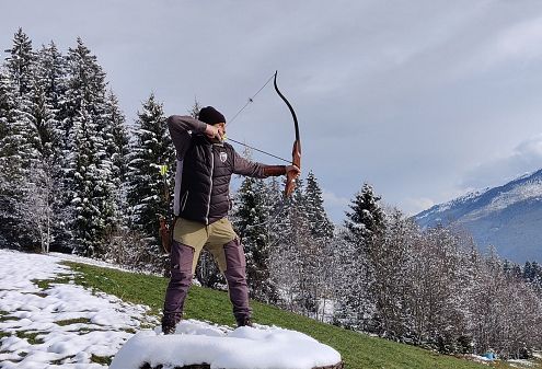 Eine Person steht auf schneebedecktem Boden und zielt mit einem Bogen. Im Hintergrund sind verschneite Bäume und Berge zu sehen. Der Himmel ist bewölkt.