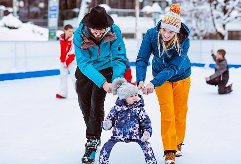 Eine lachende Familie fährt mit einem kleinen Kind auf einem Schlitten über eine Eisbahn. Der Vater schiebt, die Mutter hält fest. Im Hintergrund spielen weitere Kinder.