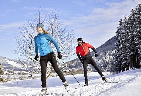 Winterlandschaft mit zwei Personen im Schnee, die Langlaufski anlegen. Die Sonne scheint über schneebedeckte Bäume und Hügel. Im Hintergrund ist eine Hütte zu sehen.