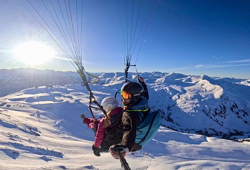 Ein Tandem-Paragliding-Duo schwebt über einer verschneiten Alpenlandschaft bei sonnigem Wetter. Der blaue Himmel und die strahlende Sonne bieten eine spektakuläre Aussicht.