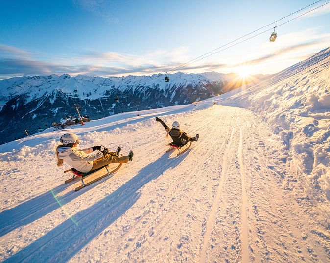 Zwei Personen rodeln bei Sonnenuntergang einen schneebedeckten Hang hinab. Im Hintergrund sind majestätische Berge und eine Seilbahn zu sehen. Der Himmel ist klar und blau.