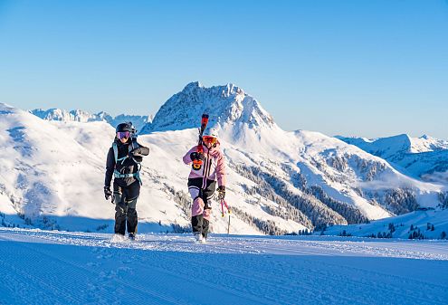 Zwei Skifahrer gehen in voller Ausrüstung über eine verschneite Berglandschaft. Sie tragen ihre Skier auf den Schultern, während die Sonne den blauen Himmel erhellt.