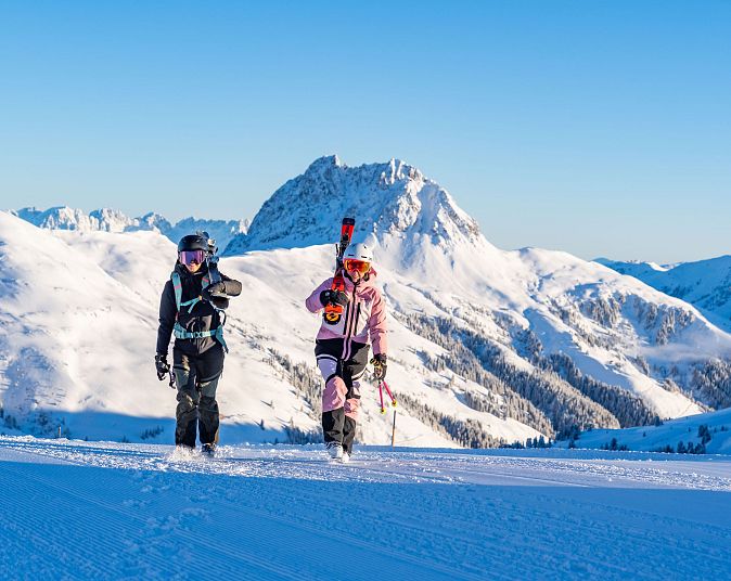 Zwei Skifahrer gehen in voller Ausrüstung über eine verschneite Berglandschaft. Sie tragen ihre Skier auf den Schultern, während die Sonne den blauen Himmel erhellt.