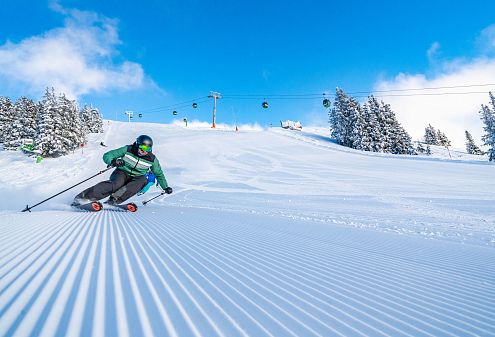 Ein Skifahrer in grüner Jacke fährt dynamisch eine frisch präparierte Skipiste hinunter. Im Hintergrund sind verschneite Bäume und eine Seilbahn vor blauem Himmel.