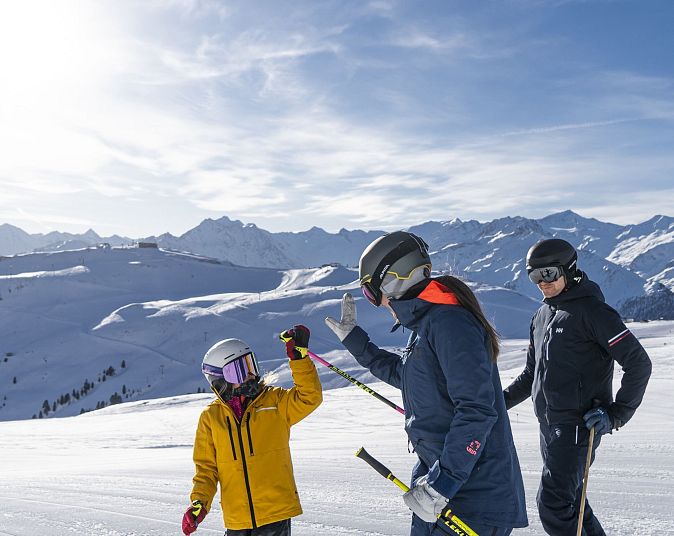 Eine Familie in Skikleidung steht auf einer schneebedeckten Skipiste. Eine Person gibt einem Kind im gelben Anzug ein High-Five. Im Hintergrund sind Berge zu sehen.