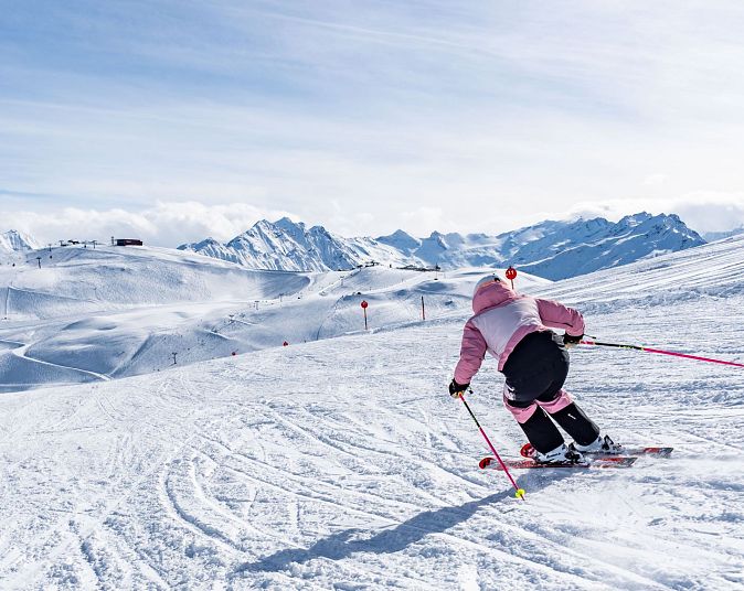 Eine Skifahrerin in rosa Kleidung fährt eine schneebedeckte Piste hinunter. Im Hintergrund sind verschneite Berge unter einem blauen Himmel zu sehen.
