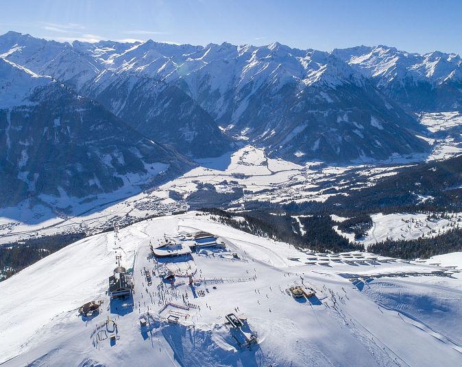 Luftaufnahme einer verschneiten Berglandschaft mit Skiliften und Pisten am Wildkogel. Im Hintergrund erstrecken sich majestätische Berggipfel unter einem klaren blauen Himmel.