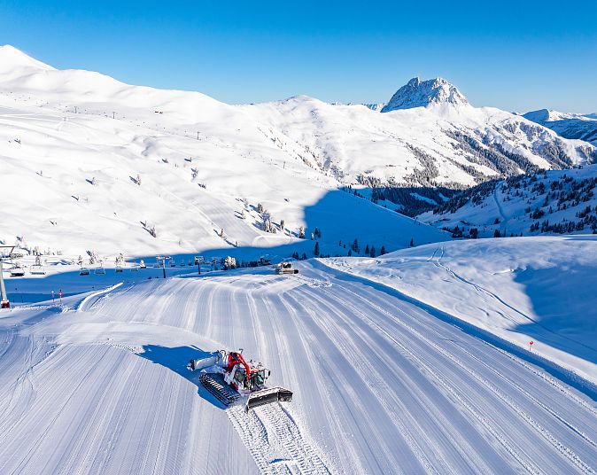 Eine Pistenraupe glättet eine breite Skipiste, umgeben von schneebedeckten Bergen unter klarem, blauem Himmel, perfekt für Wintersport bei Sonnenschein.