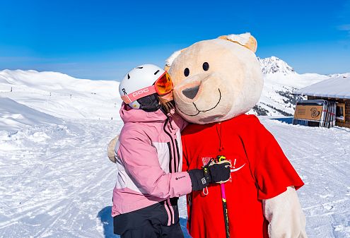 Eine Person in rosa Skijacke und Helm umarmt ein großes Teddybärkostüm in rotem Shirt auf einer schneebedeckten Piste mit blauem Himmel und Bergen im Hintergrund.