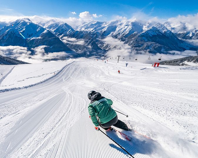 Skifahrer in der Wildkogel-Arena auf einer perfekt präparierten Piste, umgeben von verschneiten Bergen und blauem Himmel. Skilifte und weitere Skifahrer im Hintergrund.