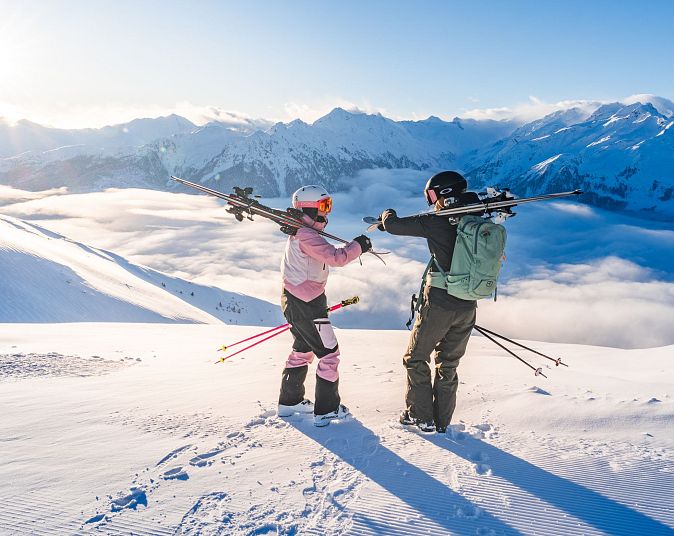 Twee mensen in skikleding staan op een besneeuwde bergtop met ski's op hun schouders en kijken uit over een prachtig berglandschap met mist en zon.