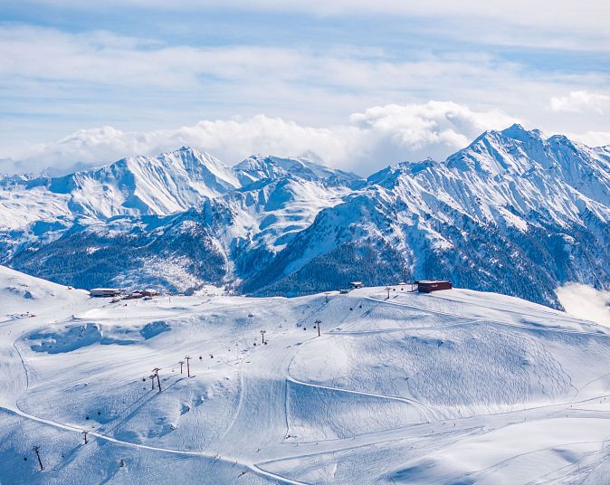 Snowy alpine landscape with ski lifts, surrounded by majestic mountain peaks under a clear blue sky with some clouds, creating a serene winter scene.