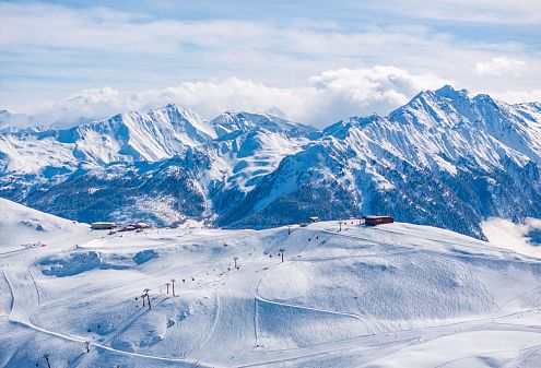 Berglandschap met besneeuwde toppen en skiliften tegen een heldere hemel. De zon schijnt fel en markeert de contouren van de bergen.