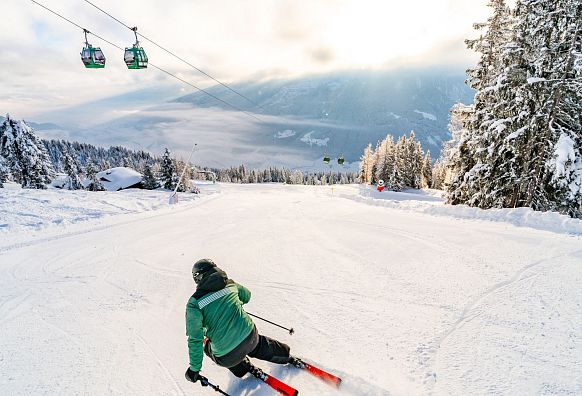 Ein Skifahrer in grüner Jacke fährt eine schneebedeckte Piste hinunter. Im Hintergrund sind Bäume, Berge und schwebende Gondeln zu sehen.