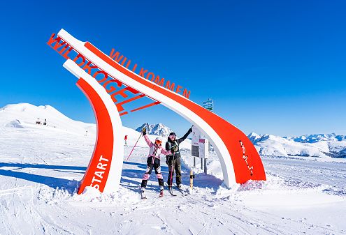 Zwei Skifahrer stehen neben einem modernen, rot-weißen Schild mit der Aufschrift "Wildkogel" im Schnee. Im Hintergrund sind verschneite Berge und blauer Himmel zu sehen.