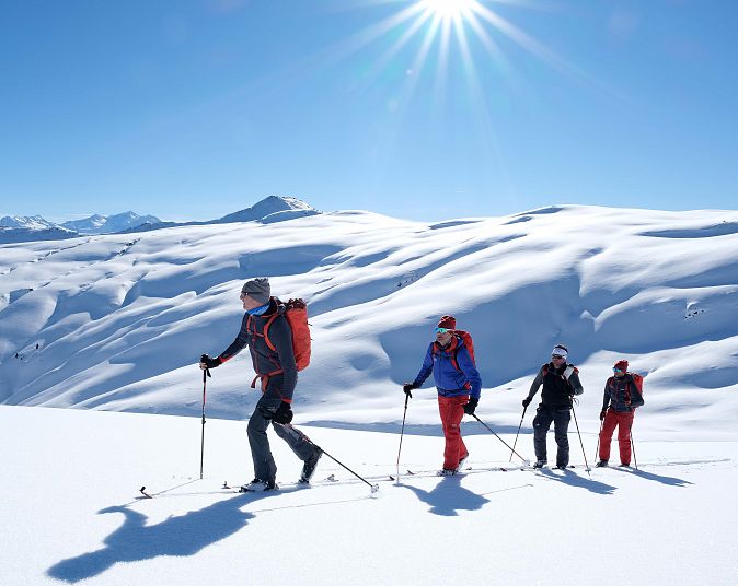 Vier Personen wandern unter strahlendem Sonnenschein mit Ski und Stöcken durch eine schneebedeckte Gebirgslandschaft. Der Himmel ist klar und die Berge weitläufig.