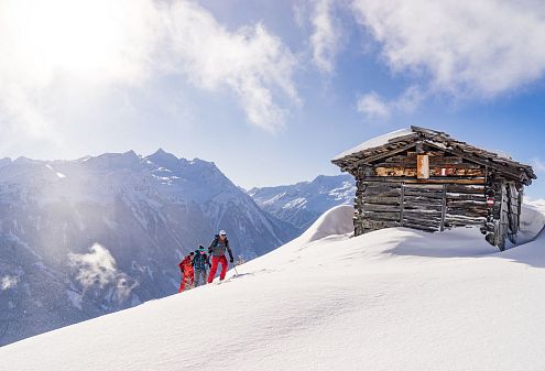 Drei Skitourengeher in Skikleidung gehen durch tiefen Schnee zu einer hölzernen Berghütte. Im Hintergrund beeindruckende verschneite Berge unter blauem Himmel.