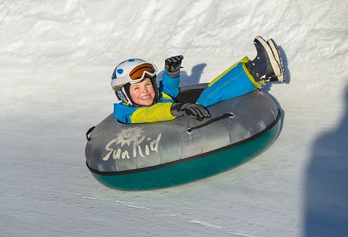 Zwei Personen rodeln mit aufblasbaren Reifen die Snowtubingbahn beim Wennsergletscher in Bramberg hinunter. Die vordere Person trägt eine schwarze Jacke, blauen Helm und weiße Schutzbrille.