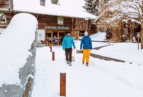 Zwei Personen spazieren im Schnee vor dem Eingang vom Museum Bramberg. Sie tragen warme Winterkleidung. Das Umfeld ist von verschneiten Bäumen und Holzhäusern geprägt.
