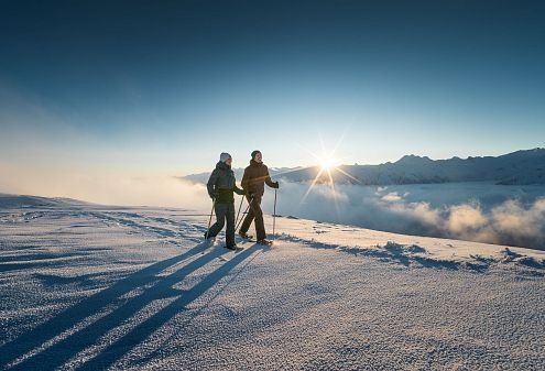 Zwei Personen stehen im Schnee auf einem Berggipfel bei Sonnenaufgang. Sie tragen Winterkleidung und Skistöcke. Der Himmel ist klar und der Horizont von Bergen gesäumt.