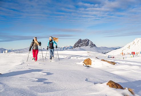 Two people cross-country skiing in a snowy landscape with a mountain backdrop and a clear blue sky. They are bundled up in winter clothing.