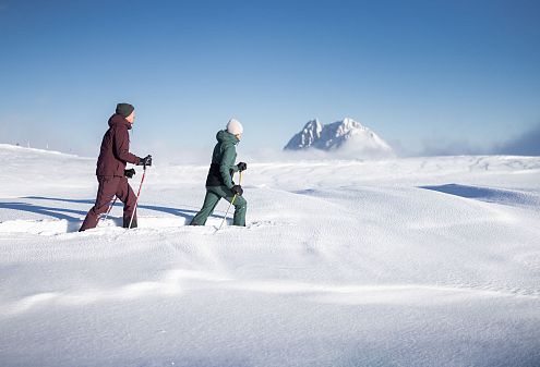 Dva lidé v zimním oblečení se procházejí zasněženou krajinou s hůlkami, na pozadí je vidět zasněžená hora pod modrou oblohou.